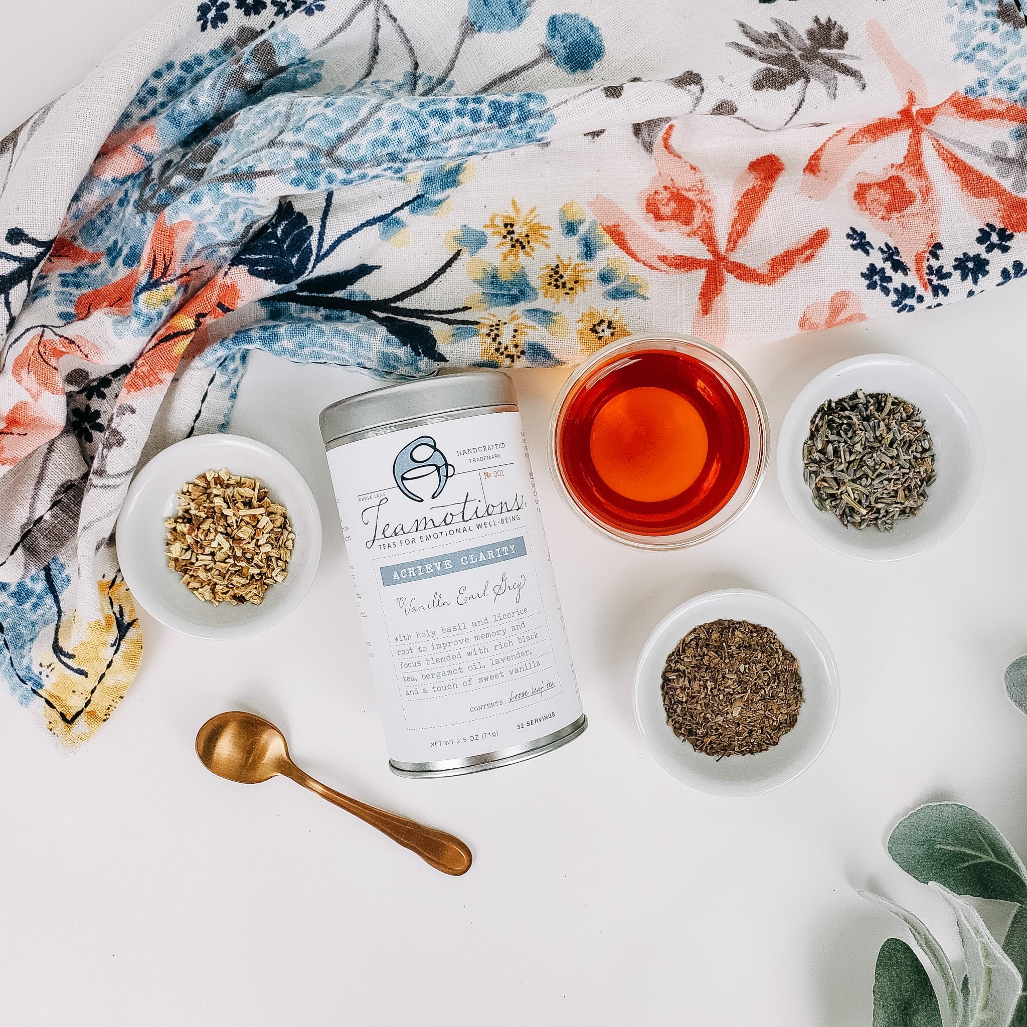 A tin of Achieve Clarity vanilla Earl Grey beside a glass of brewed tea and small bowls of holy basil, licorice root, and lavender