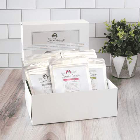 Box of assorted teas with pouches, placed on a wooden table near a plant.