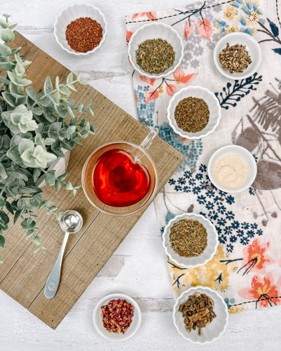 A heart-shaped glass mug of tea surrounded by herbs